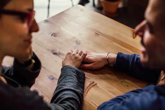 A close-up photograph of two individuals seated at a wooden table, gently holding hands, conveying a sense of support and connection.